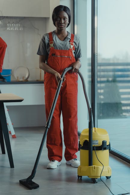 A person dressed in protective clothing, including gloves and a disposable suit, is operating a vacuum cleaner with a long hose and a flat cleaning head, performing surface cleaning on a tiled and woo
