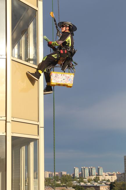 Two professional window cleaners from Hounslow Cleaners are suspended on a platform attached to a tall, modern building with a brick façade and large glass windows. They are wearing safety harnesses