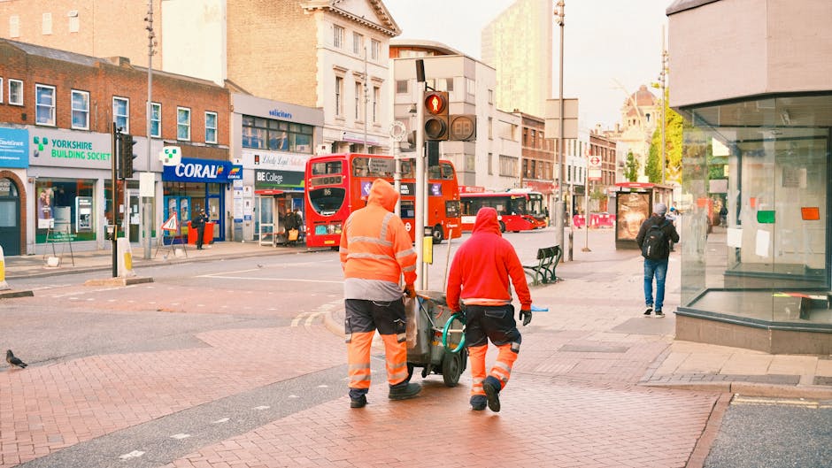 Two street cleaning workers in high-visibility orange and red uniforms are currently engaged in surface cleaning on Hounslow High Street, TW3. One worker is pushing a cleaning cart while the other pulls a hose, likely used for sanitisation or deep cleaning of the paved pedestrian area. The scene is illuminated by natural daylight, with a background of commercial buildings, including shops with glass facades and signage such as 'Coral,' and red double-decker buses traveling along the road. The surfaces, including brick-paved sidewalks and smooth tiled storefronts, appear clean and well-maintained, reflecting ongoing professional cleaning services by Hounslow Cleaners. Pedestrians are visible in the background, some walking and others waiting at nearby bus stops, contributing to a busy urban environment focused on hygiene and surface maintenance.