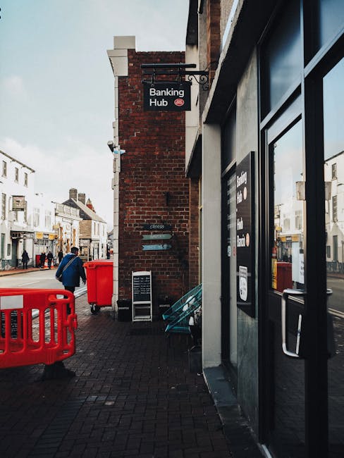Exterior view of a building on Hounslow High Street featuring a prominently displayed 'Banking Hub' sign mounted on a red brick wall. The sidewalk is paved with dark bricks, and in the foreground, there are red safety barriers and a red bin placed next to a glass door with dark framing. The scene captures a typical busy street with pedestrians dressed in winter clothing walking past white terraced buildings and shop fronts in the background. Although the image does not depict cleaning processes directly, it illustrates an urban environment maintained with clean, well-kept surfaces. This setting aligns with guidelines for surface cleaning and sanitation, reflecting a tidy, hygienic streetscape as part of regular commercial and municipal maintenance. For professional domestic cleaning and surface sanitisation services, contact Hounslow Cleaners, specialists in deep cleaning and hygiene for residential properties in TW3 and Hounslow.