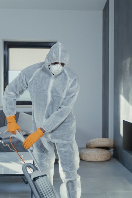 A person dressed in protective overalls, gloves, and sneakers is using a vacuum cleaner with a long hose and flat cleaning head to perform surface cleaning on a tiled floor in an indoor setting. The b